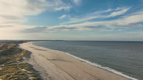 Long,-high-aerial-shot-of-a-calm-sea,-nearly-empty-beach-and-paths-through-grassy-dunes-at-sunset-on-a-beautiful-day