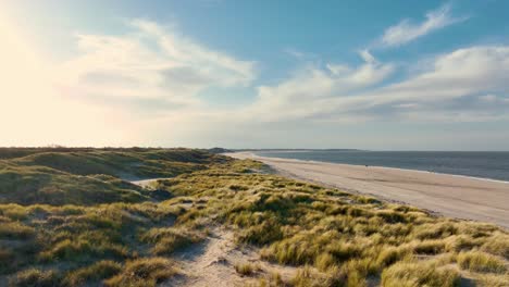 Long-aerial-shot-flying-over-grassy-dunes-along-an-empty-beach-during-a-beautiful-sunset