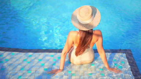 With-her-back-to-the-camera,-a-woman-sitting-on-the-shallow-step-of-a-swimming-pool-looks-around