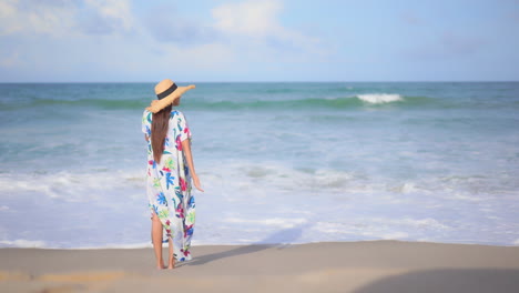 Lonely-Stylish-Female-in-Colorful-Summer-Dress-and-Hat-Standing-on-Sand-in-Front-of-Tropical-Sea-Waves,-Back-View