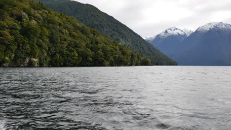Pov-boat-cruise-on-natural-lake-surrounded-by-giant-mountains-with-snow-in-Fiordland-National-park