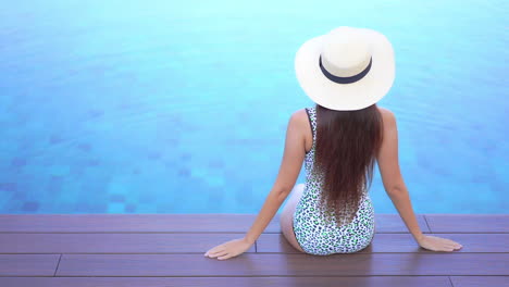 A-woman-with-her-back-to-the-camera-sits-on-the-deck-of-a-swimming-pool-with-her-feet-in-the-water