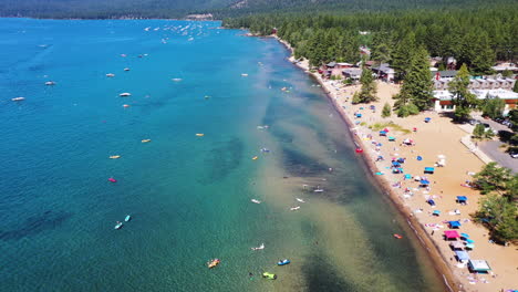 Aerial-landscape-view-of-boats-on-clear-water-and-people-relaxing-on-a-sandy-beach-surrounded-by-a-pine-forest-in-Lake-Tahoe,-Nevada
