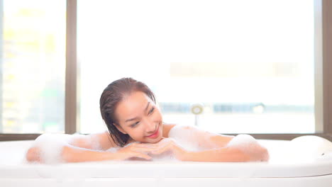 Beautiful-asian-woman-enjoying-in-hot-bathtub-full-of-foam,-sexy-female-bathing-by-the-window