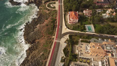 aerial-view-of-cascais-Portugal-little-city-town,-small-coastline-road-with-car-driving-along-the-country-close-to-the-rocky-ocean-cliff-beach