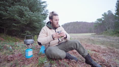 Caucasian-Guy-Sits-On-The-Ground-Next-To-A-Portable-Camper-Stove-On-Countryside-Mountains