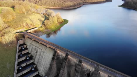 Panning-left-shot-of-a-Wimbleball-Lake-dam-and-calm-lake-reflecting-the-sky-on-a-beautiful-sunny-evening