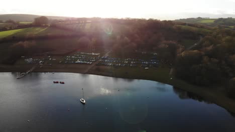 Aerial-forward-shot-of-Wimbleball-Lake-Exmoor-England-with-a-sailing-boat-moored-in-front-of-the-sailing-club-with-the-sunset-behind