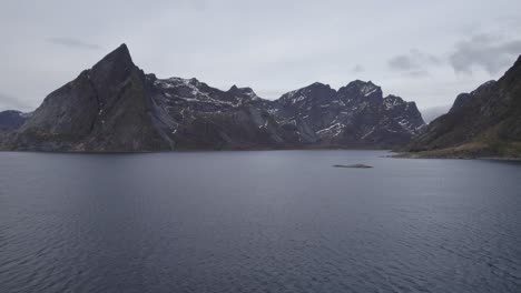 Aerial-drone-view-low-over-fjord-water-towards-steep-mountain-peaks,-in-cloudy-Lofoten,-Norway