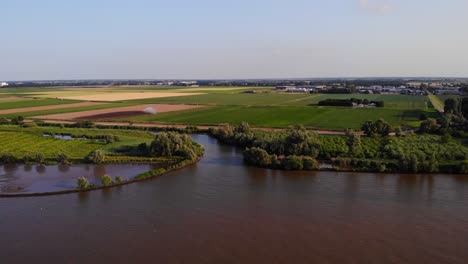 Aerial-Over-River-Waterway-Towards-Patchwork-Farmland-In-Puttershoek-With-Field-Being-Irrigated-With-Water-Sprinkler