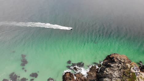 Aerial-shot-of-a-speedboat-cruising-in-turquoise-waters-and-past-headland-rocks-at-St-Ives-Cornwall-England