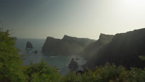 Jagged-cliffs-of-volcanic-island-with-bright-morning-sunlight,-ground-vegetation-in-foreground,-scenic-view