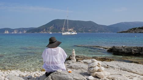 Back-View-Of-A-Girl-Sitting-On-The-Rocky-Coast-Of-Emplisi-Beach-With-Yachts-Anchored-In-The-Ocean-In-The-Background