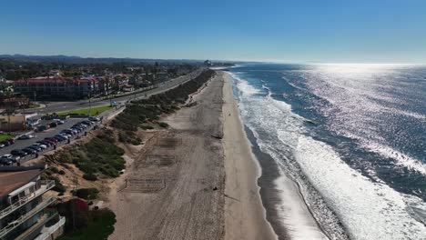 Aerial-drone-shot-over-the-sandy-beach-of-Carlsbad,-California,-USA-and-the-pacific-coast-highway-passing-by-beside-the-beach-on-a-townscape