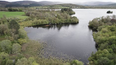 Aerial-drone-footage-flying-up-and-over-fields-and-trees-to-reveal-a-freshwater-loch-and-a-hilly,-woodland-landscape-with-rain-in-the-distance