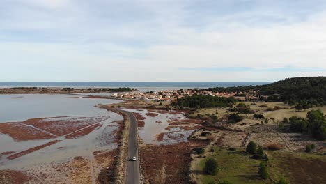 Aerial:-car-going-through-a-flat-road-among-a-marsh-and-heading-for-a-coastal-town-by-the-mediterranean-sea-in-southern-France