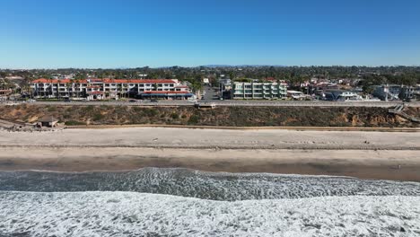 Aerial-backward-moving-shot-over-the-pacific-ocean-with-the-view-of-the-sandy-beach-and-the-town-of-Carlsbad-California,-USA-with-waves-crashing-on-the-beach