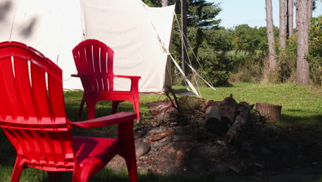 Zwei-Stühle-Stehen-Tagsüber-In-Der-Nähe-Eines-Lagerfeuers-Im-Wald,-Während-Im-Hintergrund-Ein-Zelt-Aufgebaut-Wird