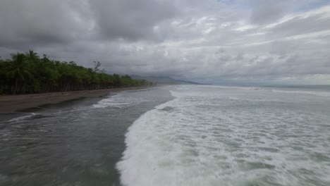 Aerial-dolly-in-over-foamy-sea-waves-hitting-the-sand-near-dense-jungle-on-a-cloudy-day-in-Dominicalito-Beach,-Costa-Rica