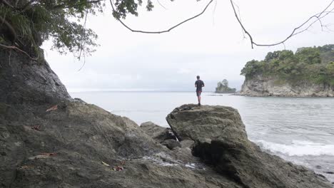 Young-man-standing-on-the-rocky-shoreline-of-Playa-La-Vaca,-a-tropical-beach-near-Quepos,-Costa-Rica