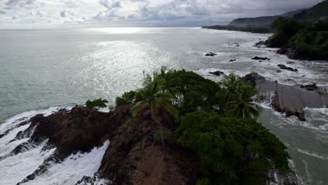 Aerial-orbit-of-rocky-shore-covered-in-vegetation-and-foamy-turquoise-sea-waves-in-in-Dominicalito-Beach,-Costa-Rica