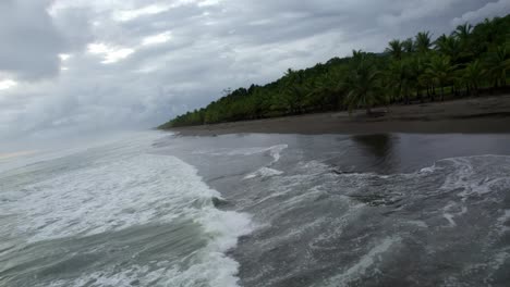 Aerial-dolly-in-of-foamy-sea-waves-hitting-the-shore-near-jungle-vegetation-on-overcast-day-in-Dominicalito-Beach,-Costa-Rica