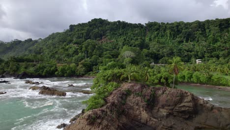 Aerial-dolly-in-of-turquoise-sea-near-rocky-shore-with-green-jungle-hillside-on-overcast-day,-Dominicalito-Beach,-Costa-Rica