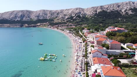 Baška-Beach,-Krk-island,-Croatia---Aerial-Drone-View-of-Tourists,-Boulevard,-Sunbeds-and-Water-Playground-at-a-Sunny-Summer-Day