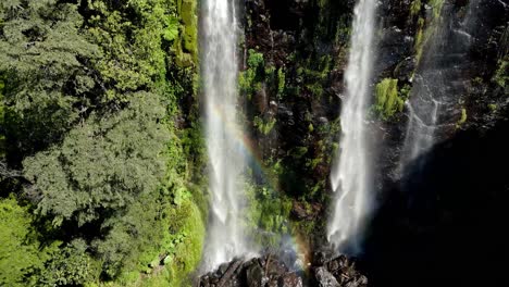 Double-waterfall-with-a-rainbow-hidden-in-a-forest---drone-shot