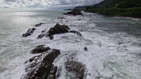 Aerial-dolly-in-over-rugged-shore-and-foamy-sea-waves-near-dense-forest-hillside-in-Dominicalito-Beach,-Costa-Rica