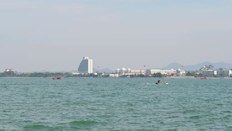 Ein-Kleines-Grünes-Fischerboot,-Das-Nach-Rechts-Fährt,-Und-Einige,-Die-In-Der-Nähe-Verankert-Sind,-Und-Die-Stadt-Pattaya-Im-Hintergrund-In-Chonburi,-Thailand