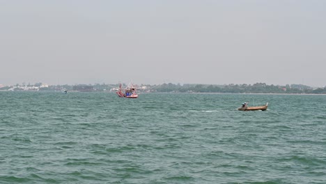 A-brown-small-fishing-boat-going-to-the-right-while-another-is-anchored-as-the-city-of-Pattaya-is-at-the-background,-Chonburi,-Thailand