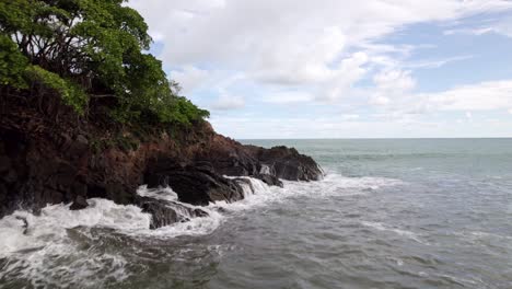 Aerial-dolly-in-over-sea-waves-hitting-the-rocky-irregular-shore-on-a-cloudy-day-in-Dominicalito-Beach,-Costa-Rica