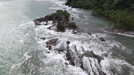 Aerial-dolly-in-over-rocky-coastline-and-turquoise-foamy-waves-near-forest-hillside-in-Dominicalito-Beach,-Costa-Rica