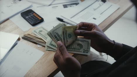 Close-up-of-man's-hand-counting-money-and-making-expense-budget-for-the-month-sitting-in-his-office-desk-with-charts-and-calculator,-money-financial-management-concept