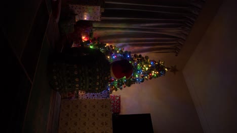 A-vertical-clip-of-a-young-man-sitting-in-front-of-a-gorgeous,-fully-lit-and-decorated-Christmas-tree,-investivating-the-presents-and-decorations-as-a-young-child-would-do-before-sitting-and-observing