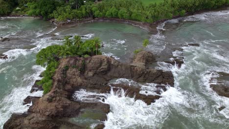 Aerial-truck-left-of-rocky-irregular-shore-and-turquoise-foamy-sea-waves-near-green-forest-in-Dominicalito-Beach,-Costa-Rica