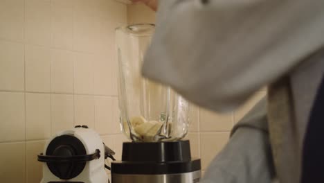 Handheld-close-up-of-young-man-putting-cut-banana-into-blender-while-making-smoothie-in-kitchen