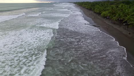Aerial-dolly-in-flying-over-turquoise-foamy-sea-waves-hitting-the-coast-near-dense-jungle-in-Dominicalito-Beach,-Costa-Rica