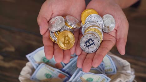 Closeup-of-man's-hand-holding-physical-bitcoin-cryptocurrency-on-brown-table-background-with-hundred-dollar-bills