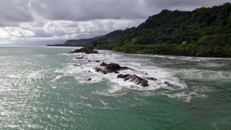 Aerial-orbit-of-turquoise-sea-near-rocky-shore-and-forest-hillside-on-a-cloudy-day-in-Dominicalito-Beach,-Costa-Rica