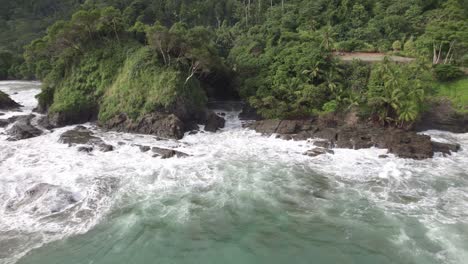 Aerial-dolly-out-rising-on-foamy-turquoise-sea-near-rocky-shore-and-green-forest-hillside-in-Dominicalito-Beach,-Costa-Rica