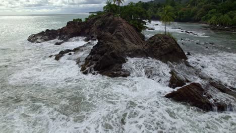 Aerial-dolly-in-flying-over-foamy-sea-waves-hitting-rocky-coastline-near-forest-hillside-in-Dominicalito-Beach,-Costa-Rica