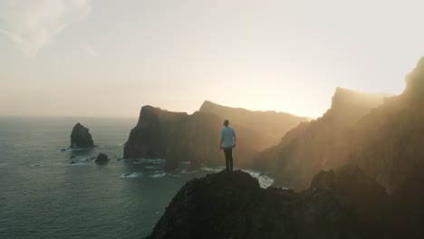 Young-male-adventure-hiker-reaching-top-of-wild-viewpoint,-looking-at-rugged-coastal-cliffs-with-bright-morning-sunlight,-successful-summit,-aerial