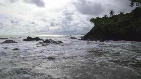 Aerial-dolly-in-flying-over-sea-waves-hitting-the-rocky-coastline-on-a-cloudy-day-in-Dominicalito-Beach,-Costa-Rica