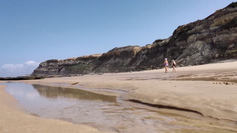 Two-elderly-lady´s-in-swimwear-walking-on-beach-near-Stone-cliffs,-Low-angle-water-stream