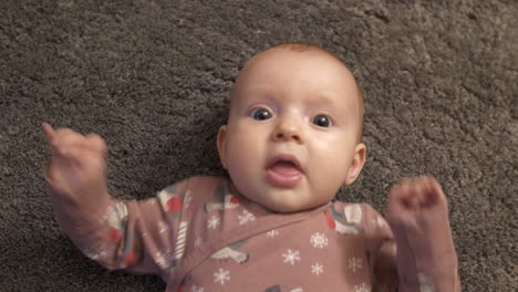 Beautiful-baby-girl-lying-on-soft-carpet-and-watches-towards-camera,-close-up-top-down-view