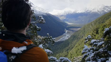 Man-hiking-on-an-adventure-through-snowy-mountains-looking-out-over-a-beautiful-green-valley-with-a-river-running-through