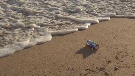 Empty-can-or-tin-on-sand-beach-and-water-foam-from-sea-coming,-environmental-pollution-concept