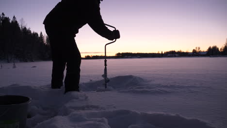 Man-drills-through-snowy-ice-to-prepare-for-ice-fishing-at-dusk
